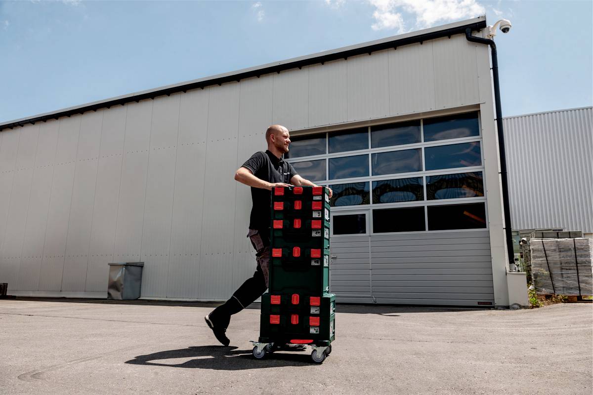 A man is pushing a trolley with stacked boxes in front of a warehouse. The sky is partly cloudy.