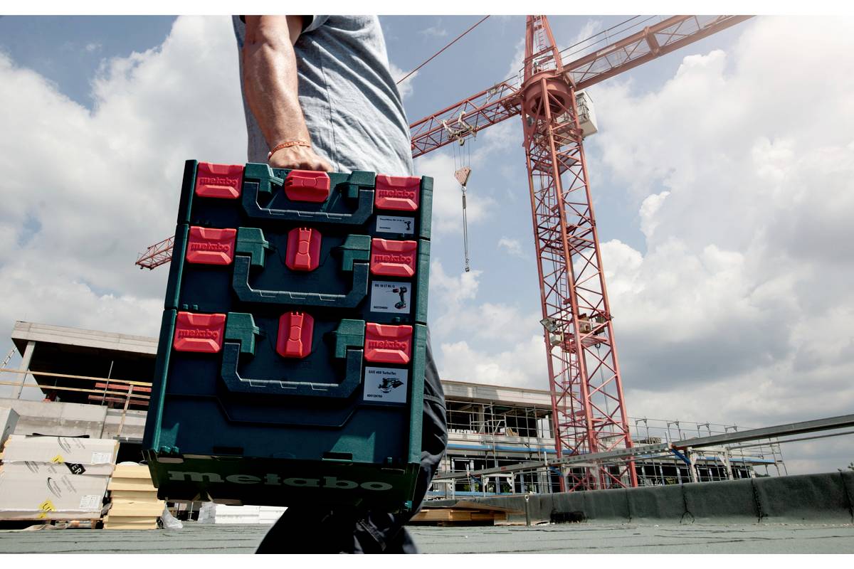 A person is carrying a toolbox on a building site. A construction crane is visible in the background. The sky is cloudy.