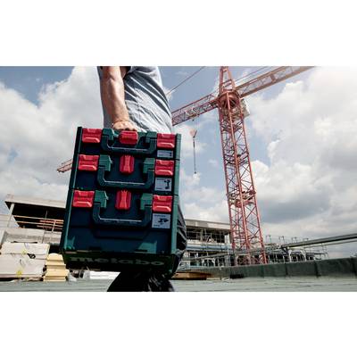 A person is carrying a toolbox on a building site. A construction crane is visible in the background. The sky is cloudy.