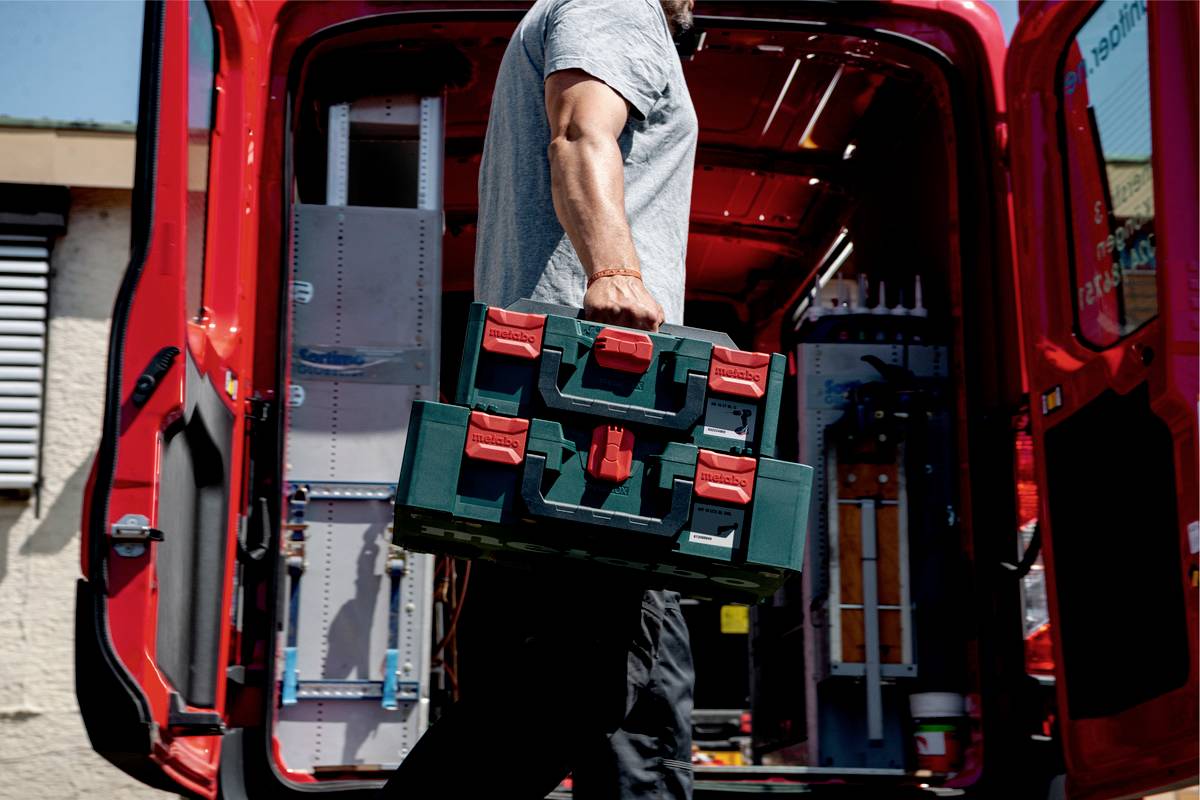 A person is carrying a toolbox into a red delivery van. Tools and shelves are visible in the background of the van.