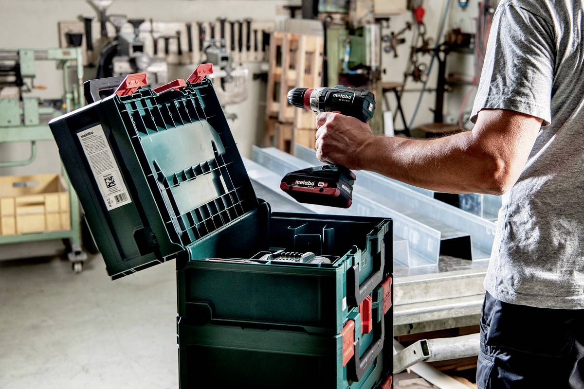 A person in a workshop is holding a cordless drill over an open toolbox, surrounded by machines and wooden furniture.