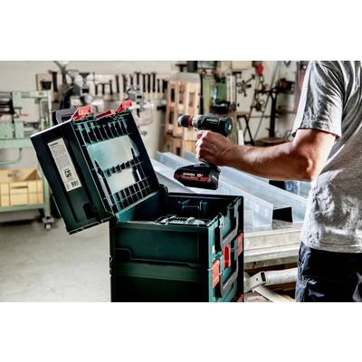 A person in a workshop is holding a cordless drill over an open toolbox, surrounded by machines and wooden furniture.