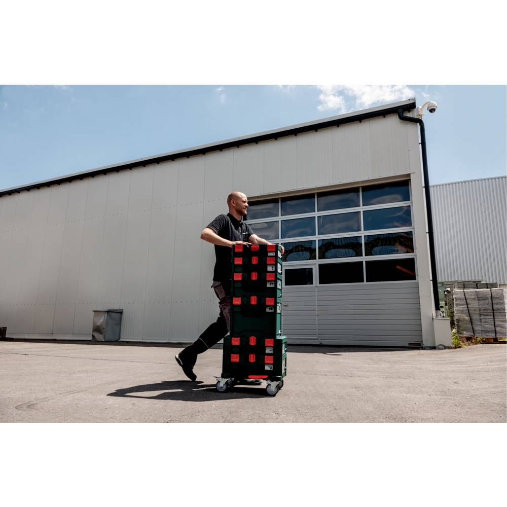 A person moves stacked crates on a cart outside a large industrial building under a clear sky, suggesting a work or delivery context.