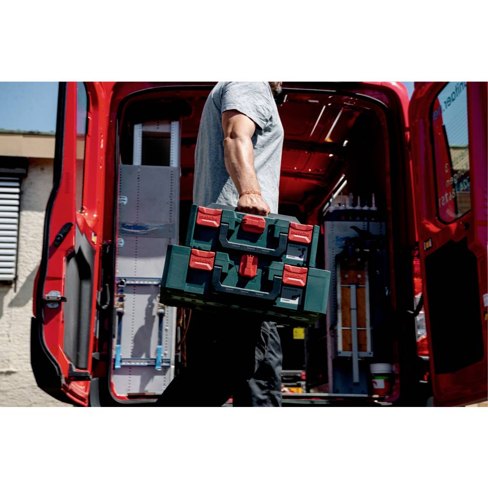 A person carrying a green and red toolbox is walking towards an open van with tool shelves inside on a sunny day.
