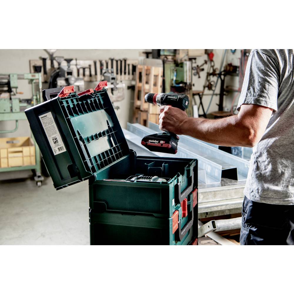 'A person holding a power drill in a workshop, using a tool box to organize equipment, with machinery visible in the background.'