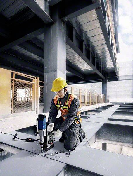 A construction worker in a hard hat and high-visibility vest kneels on a steel girder and uses a drill on a building site.