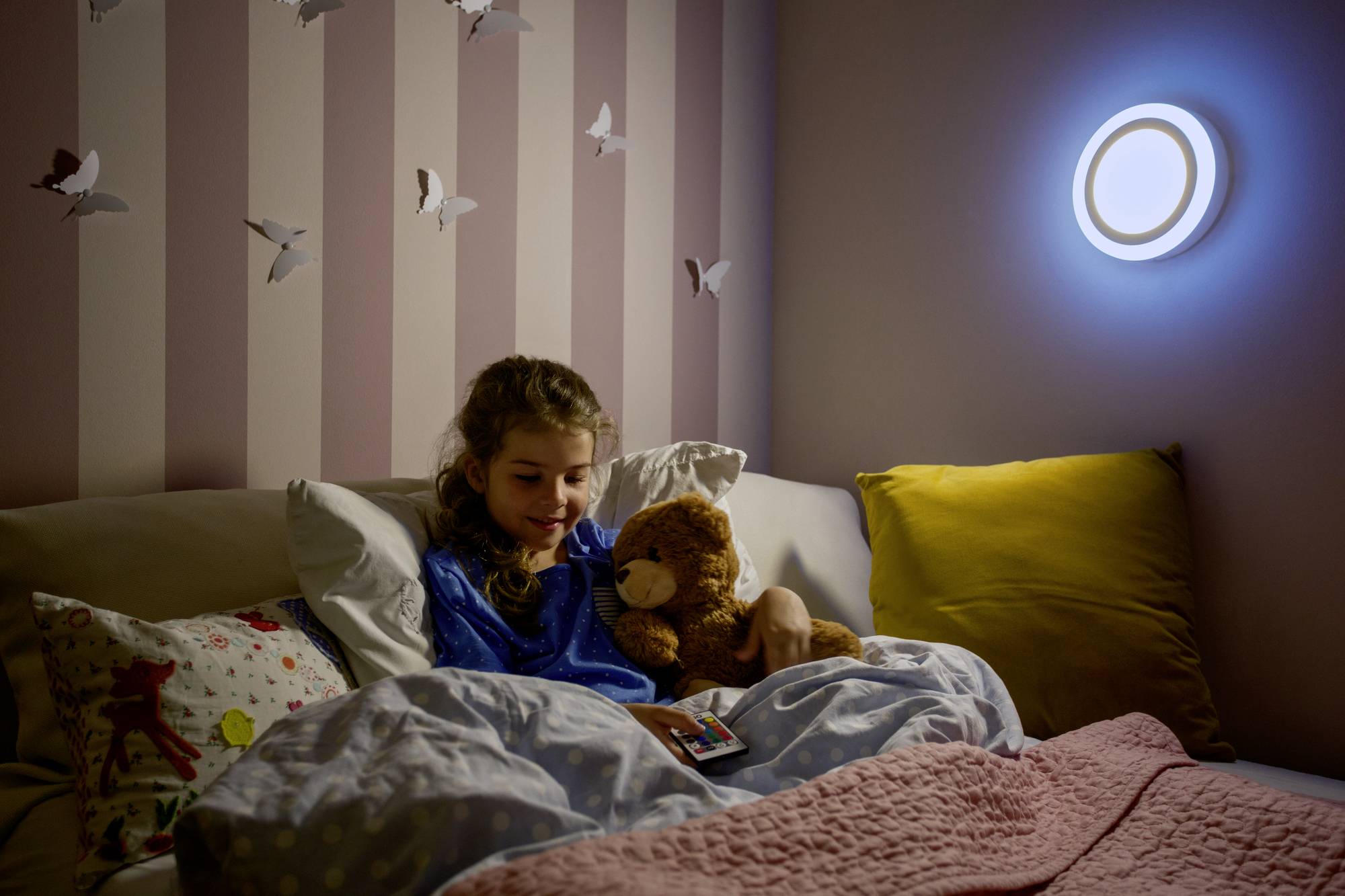 A child in pyjamas lies in bed, holding a teddy bear and looking at a book. The wall is decorated with butterflies, and a lamp is illuminating the room.