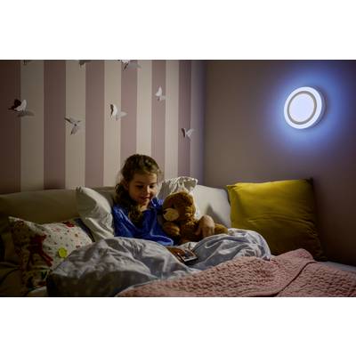 A child in pyjamas lies in bed, holding a teddy bear and looking at a book. The wall is decorated with butterflies, and a lamp is illuminating the room.