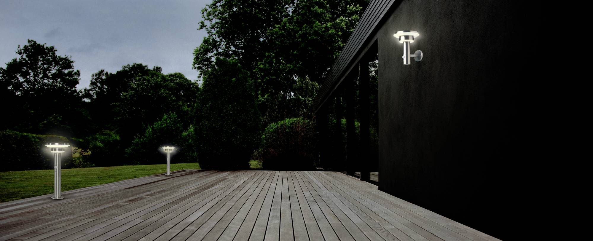A modern garden at night with a wooden deck, illuminated by two slender outdoor lights on the left and one on the wall to the right.