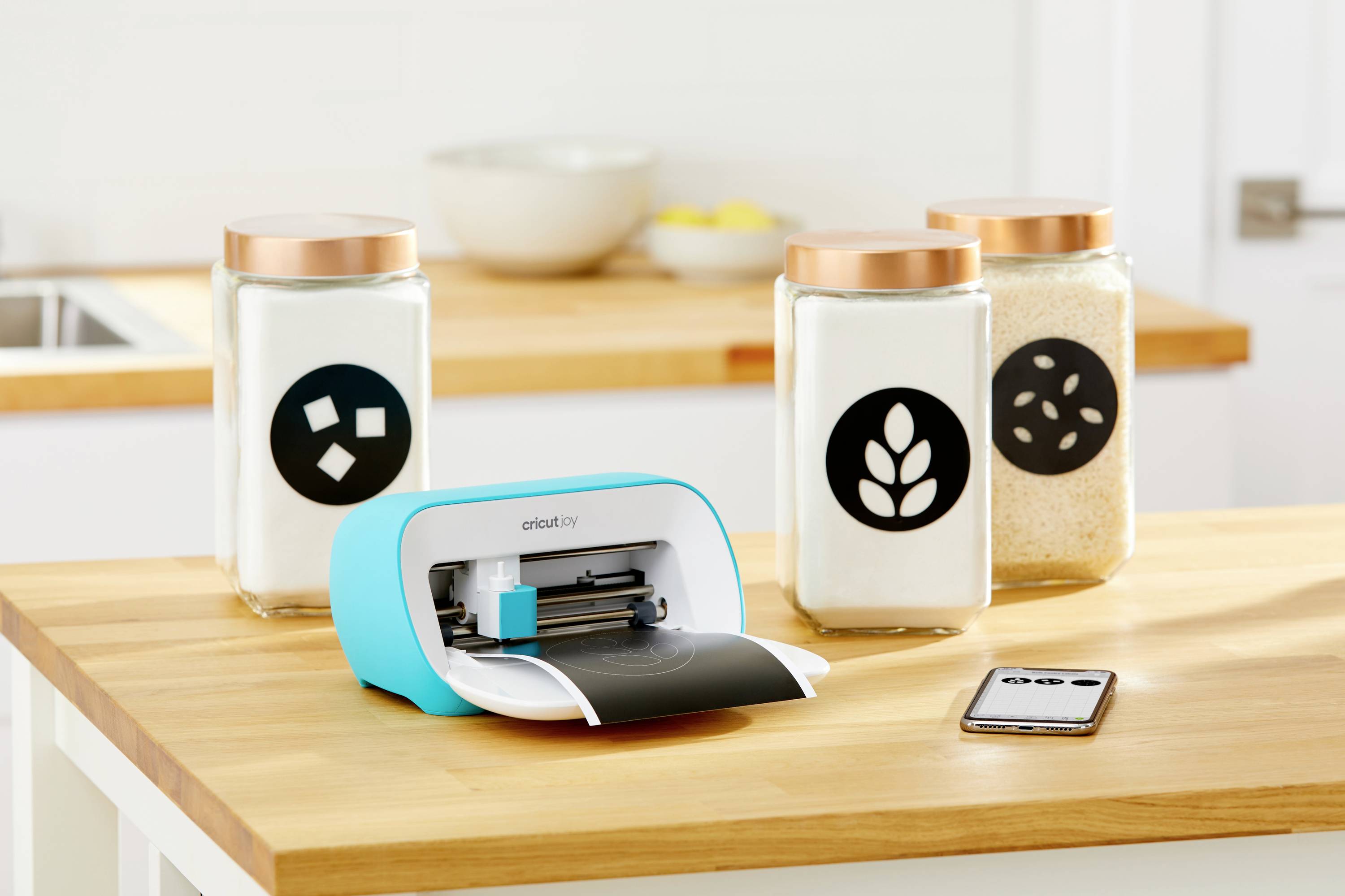 A blue cutting device on a wooden table creates labels for storage containers in the kitchen. A smartphone lies beside it.