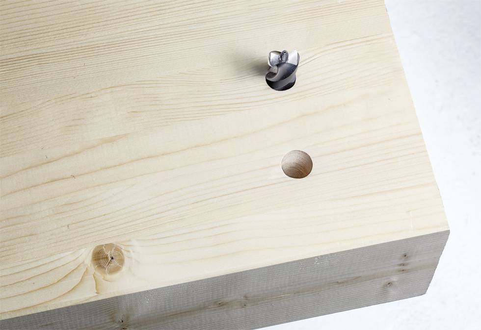 A wooden block with a drilled hole and a drill bit hovering above it. The drill bit has a spiral design.