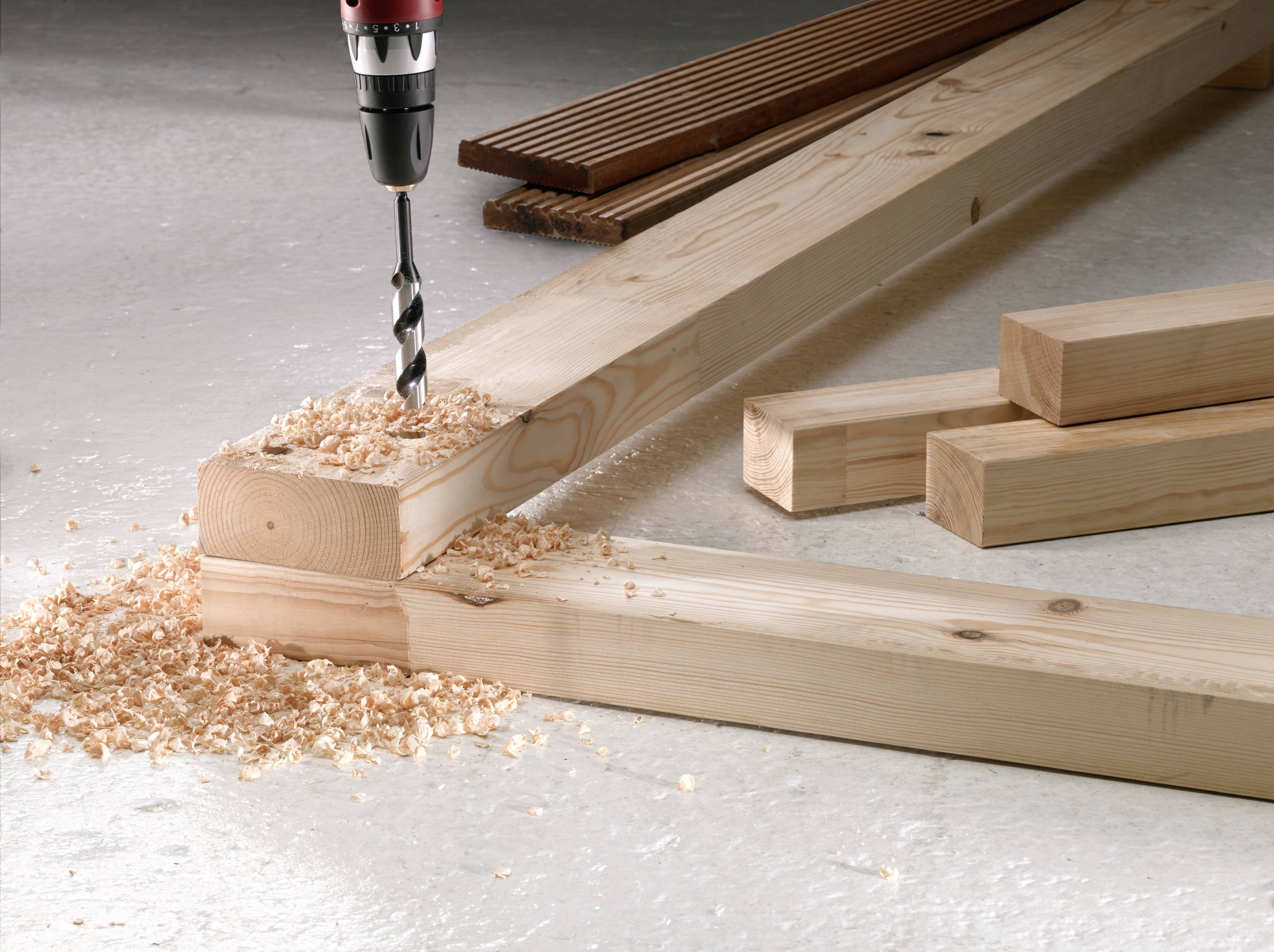 A drill is boring a hole into a wooden beam, surrounded by sawdust. Other wooden beams are lying nearby.