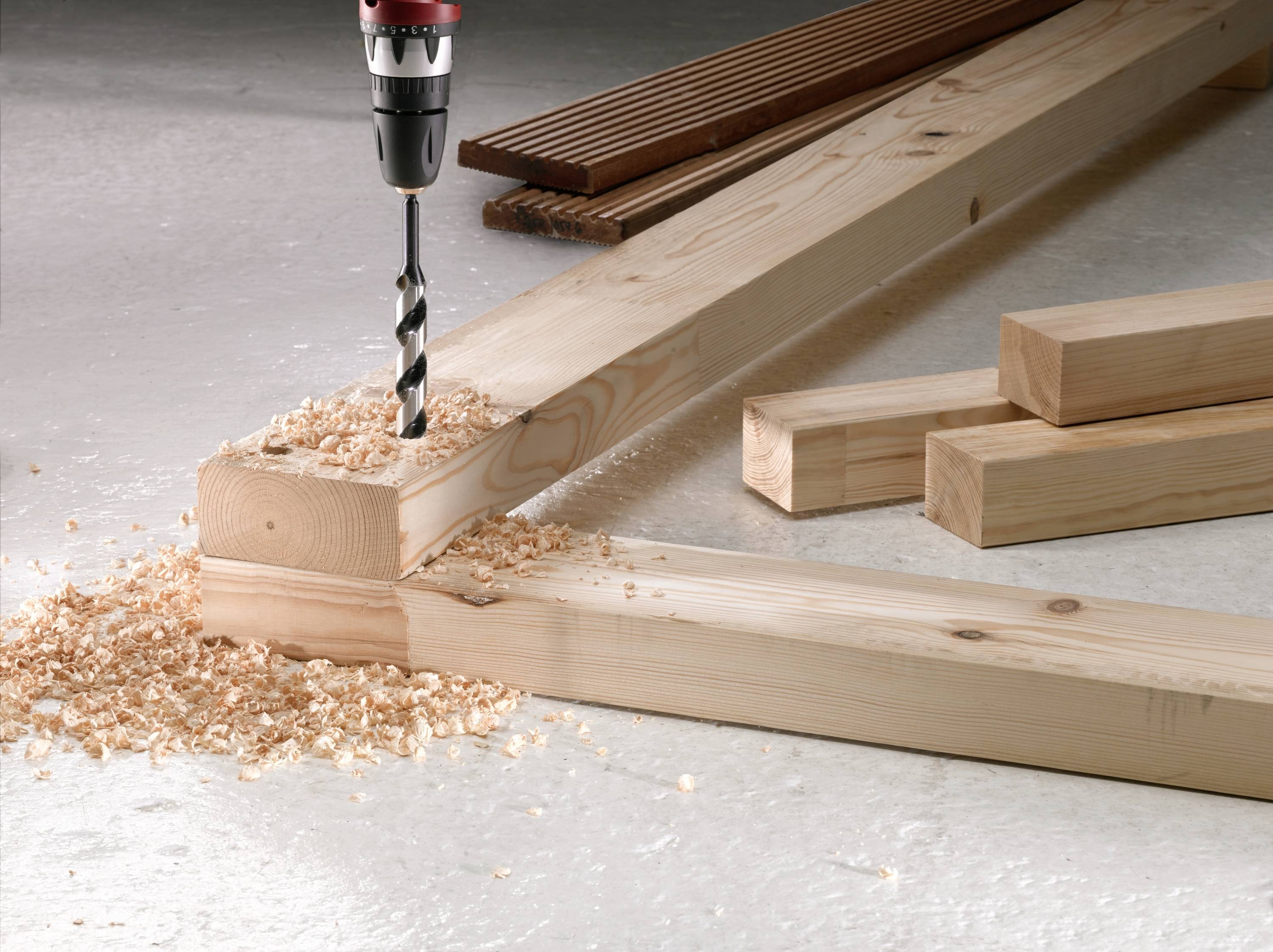 A woodworking drill bores into a wooden beam, surrounded by wood shavings. In the background, additional wooden planks are lying.