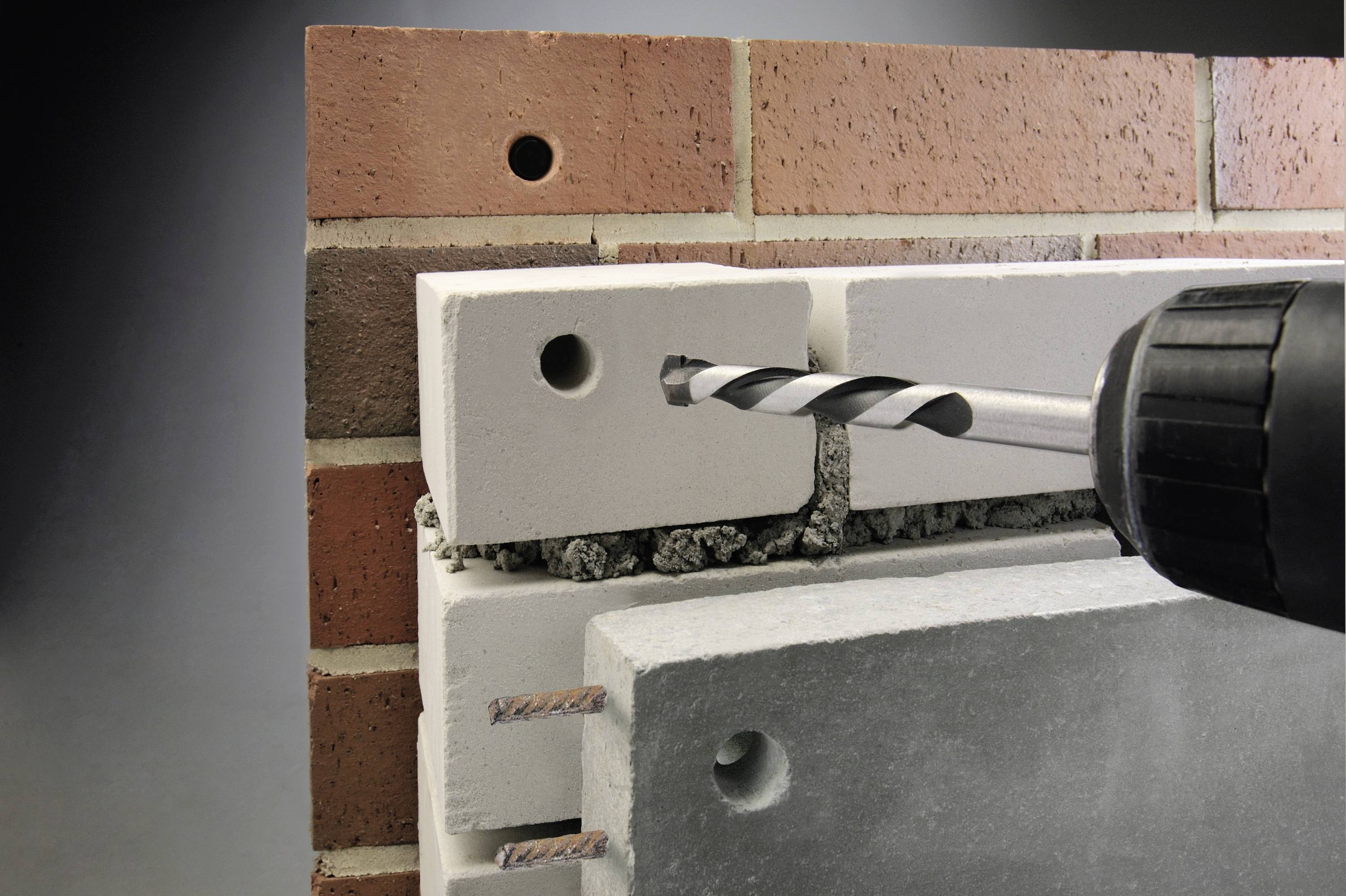 A drill is guided through a multi-layered wall composed of bricks and concrete. The layers are visibly distinct.