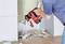 A person is using a red drill to remove tiles from a wall. Tile fragments are scattered on the floor.