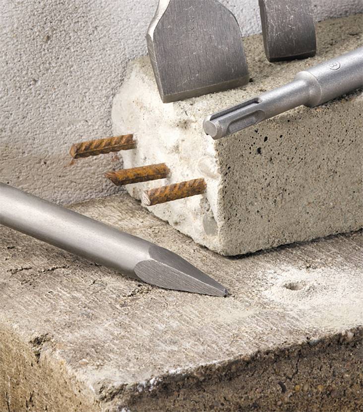 A masonry drill bit penetrating concrete and metal within a wall structure, with concrete fragments on a construction site.