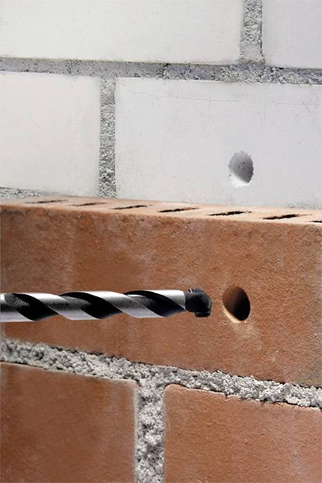 A brick with a drilled hole and a white stone lying above it with a hole. A drill protrudes from the brick's hole.