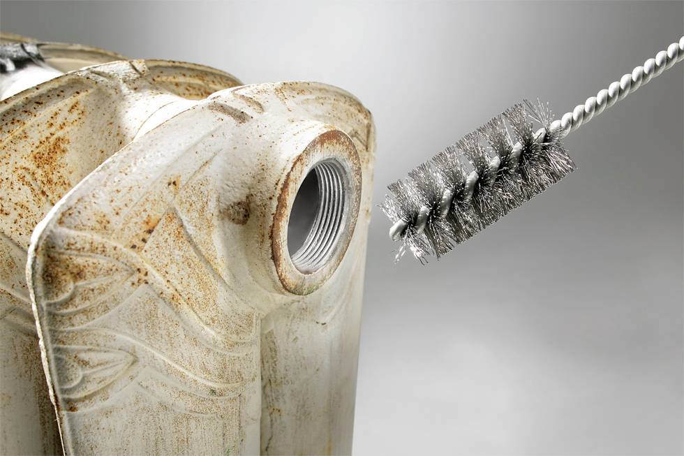 A rusted metal jerry can is being cleaned with a metal brush. The brush is positioned near the opening of the container.