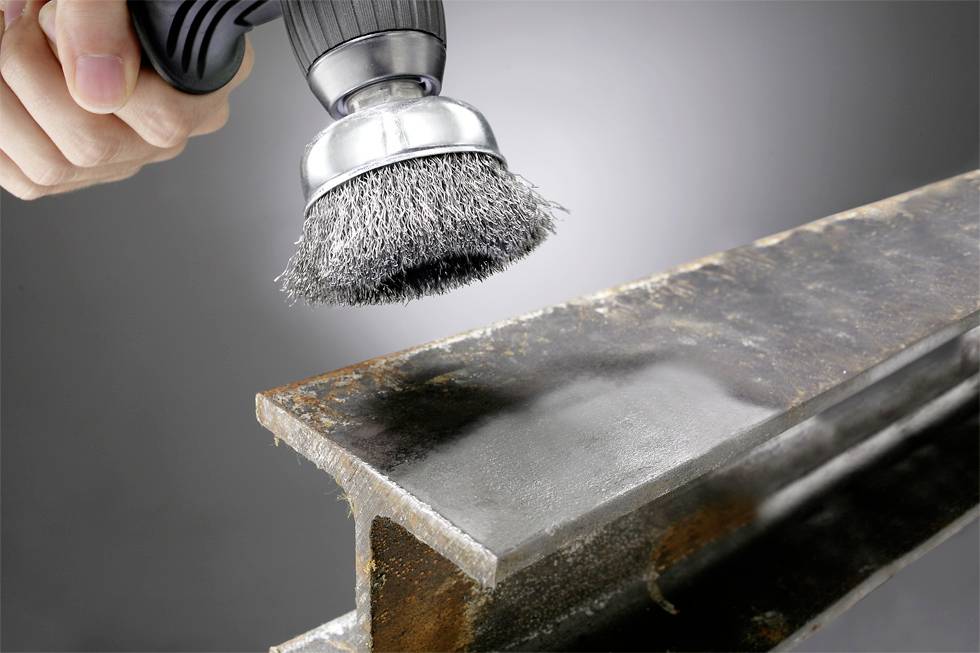 Close-up of a hand holding a wire brush on an angle grinder, removing rust from a metal beam.