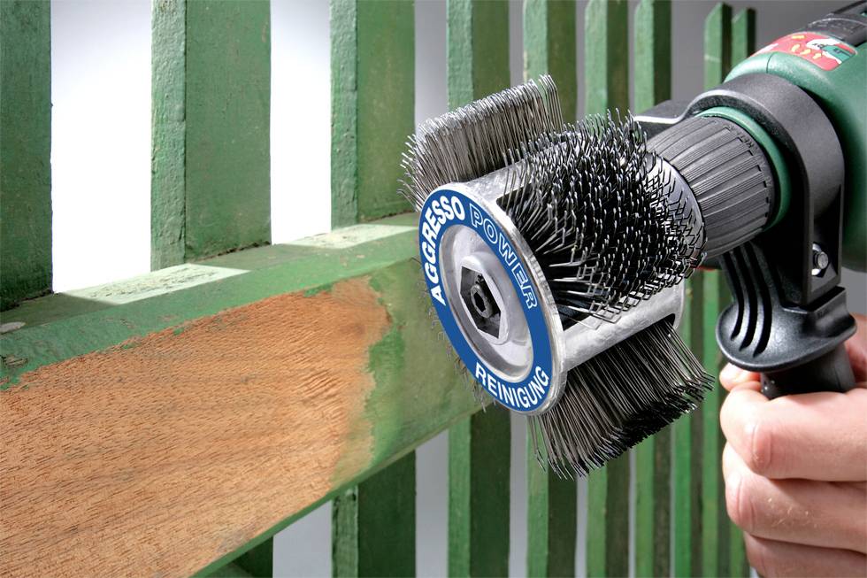 A hand holds a rotating wire brush, removing green paint from a wooden banister and exposing the wood beneath.