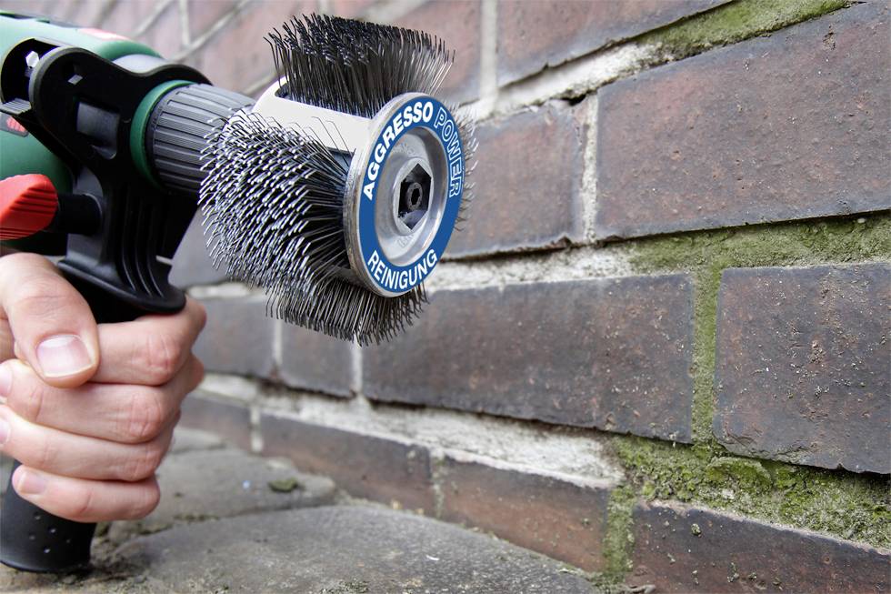 A hand is holding an electric wire brush tool that is removing mortar and deposits from the joints between brickwork.