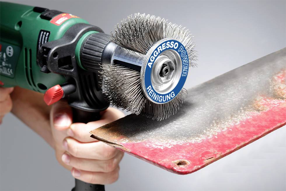 'A hand holds an electric wire brush removing rust from a metal plate. The brush is labelled "Aggresso Power Cleaning".'