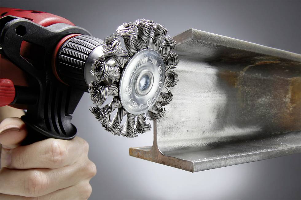 A hand holds a cordless drill with a wire brush attachment, removing rust from a metal support.