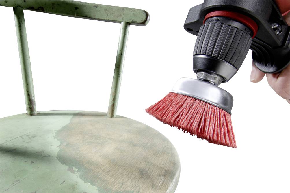 A green, flaking metal surface of a chair is being cleaned by a rotating wire brush.