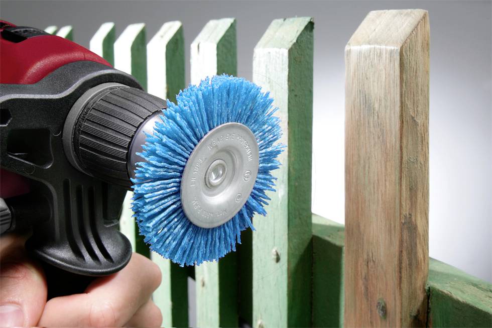 A hand-held drill driver with a blue brush is being used to clean or polish a green wooden fence.