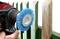 A hand is holding an electric tool with a blue wire brush to remove yellow paint from a green wooden fence.