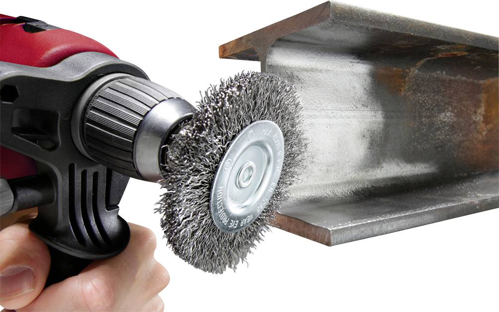 A hand is holding a drill with a wire brush attachment, cleaning rusty steel. The steel beam is partially rust-free.