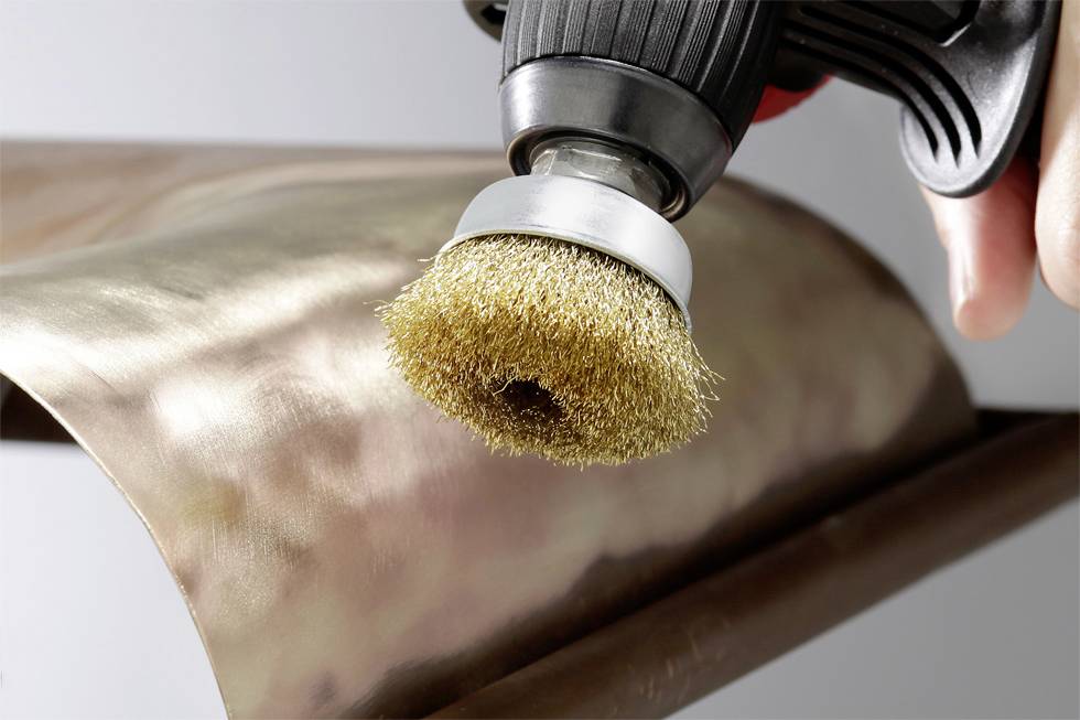 'Electric drill with wire brush attachment polishes copper metal surface, focus on brush head.'