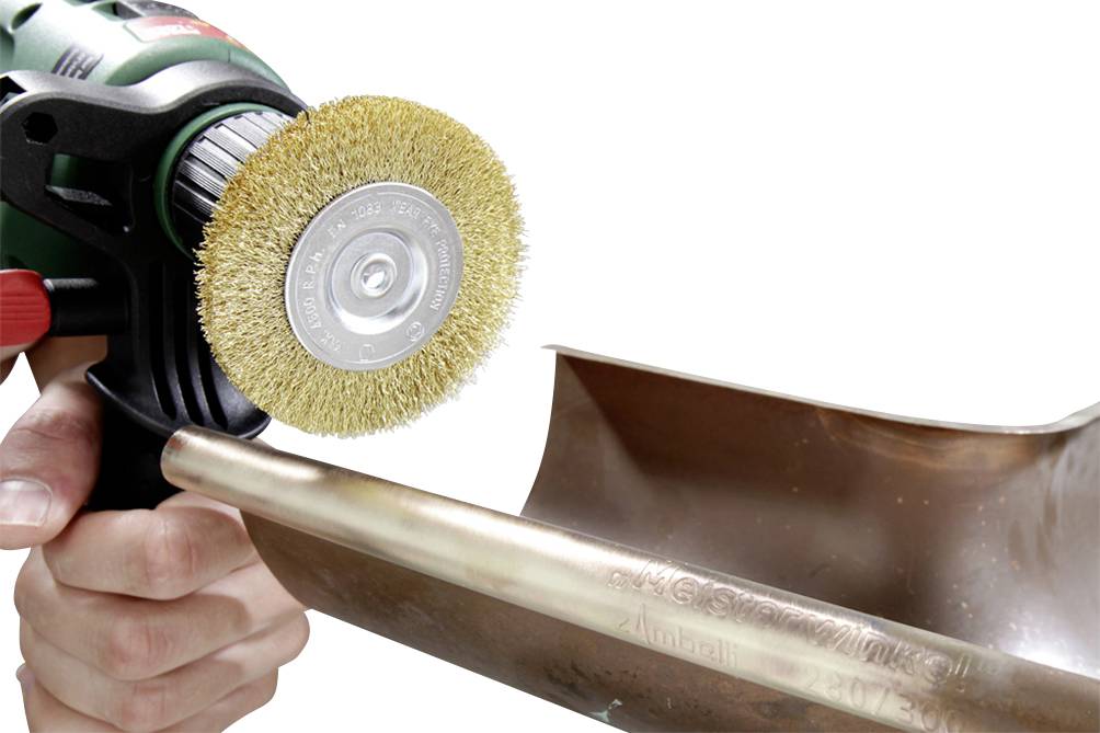 A hand holds a rotating wire brush polishing a brass pipe connection.