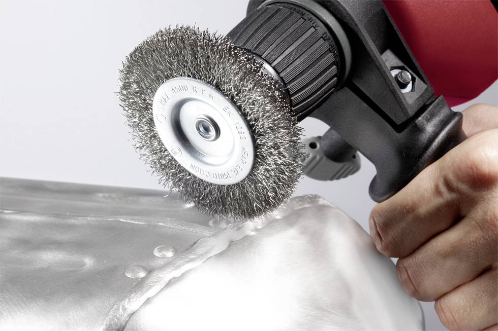 A hand is holding an electric tool with a wire brush, polishing a metal surface. The focus is on the handling of the tool.
