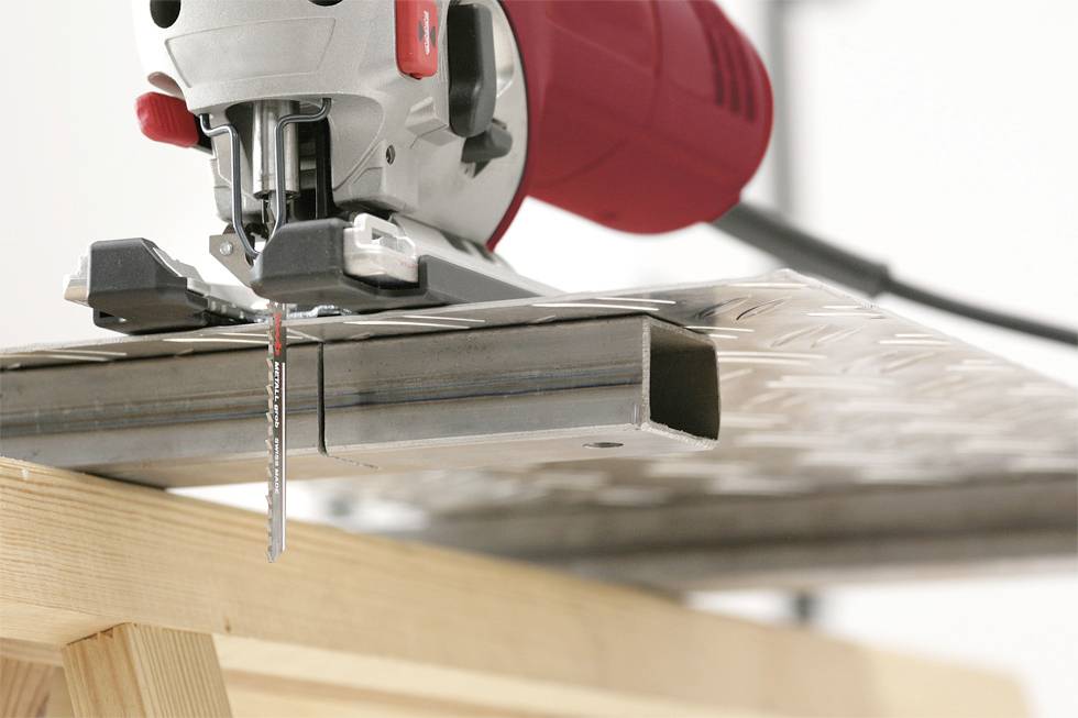 A close-up of an electric jigsaw cutting through a metal sheet; the focus is on the blade and the cutting process.