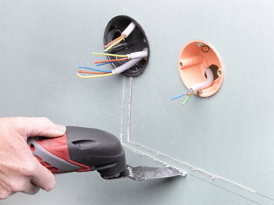 The image shows a hand using an oscillating tool to cut openings in a plasterboard sheet next to two round cable boxes.