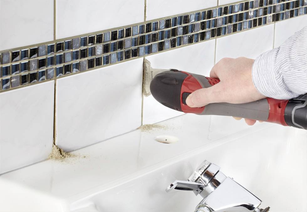 A person is using an electric tool to clean the grout between bathroom tiles above a sink.