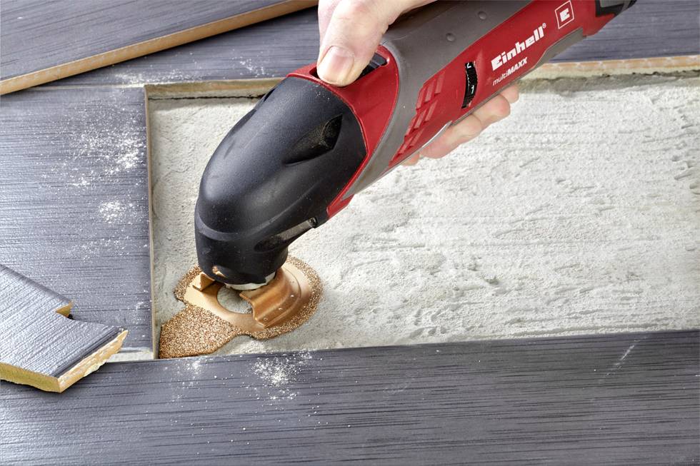 A hand is holding a red and black multi-functional tool with a circular saw that is helping to cut a section of a dark tile.