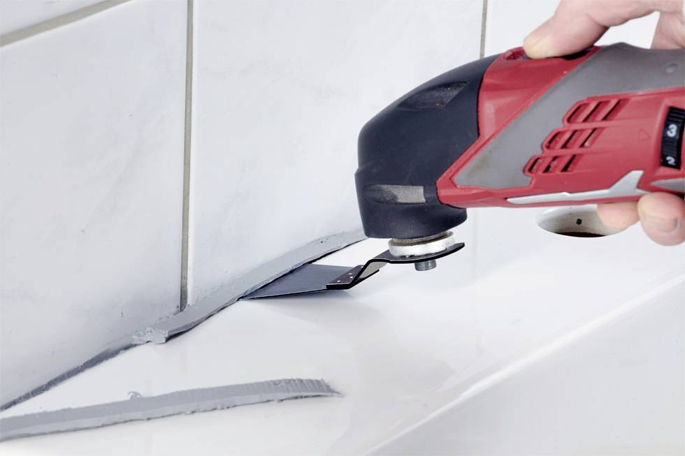 A person is using a red and black multi-tool to work on joints between white tiles and a surface.