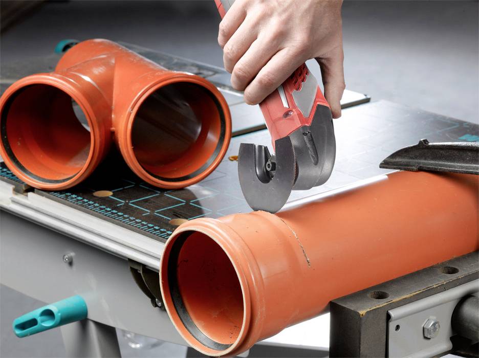 A hand is cutting an orange plastic pipe with a pipe cutter on a work bench. A further T-piece is lying beside it.