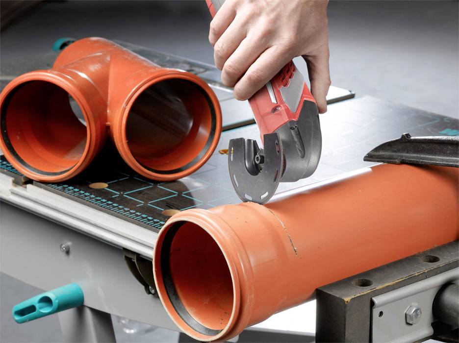 A person is cutting an orange plastic pipe on a table using a pipe cutting tool, with other pipes lying nearby.