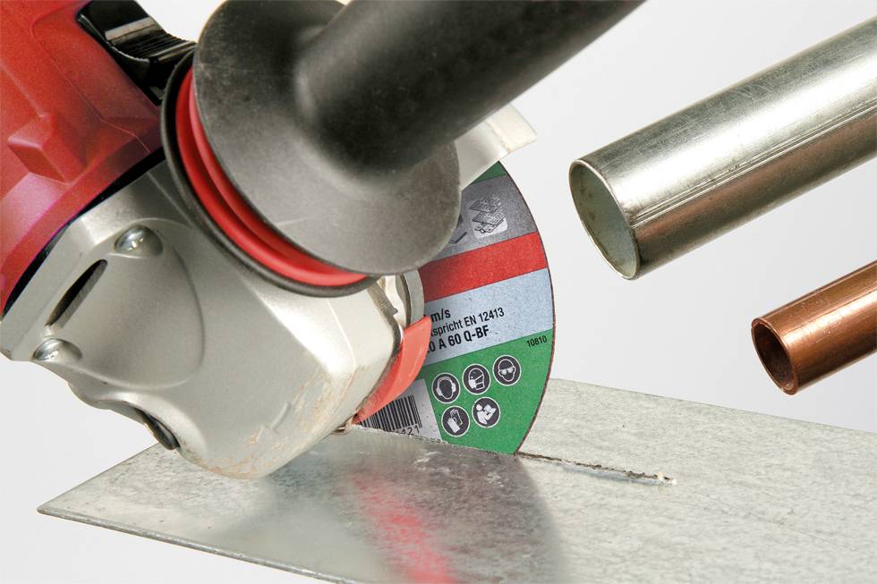 An angle grinder cuts through a metal sheet. Metal pipes are visible in the background. Focus on the cutting action.