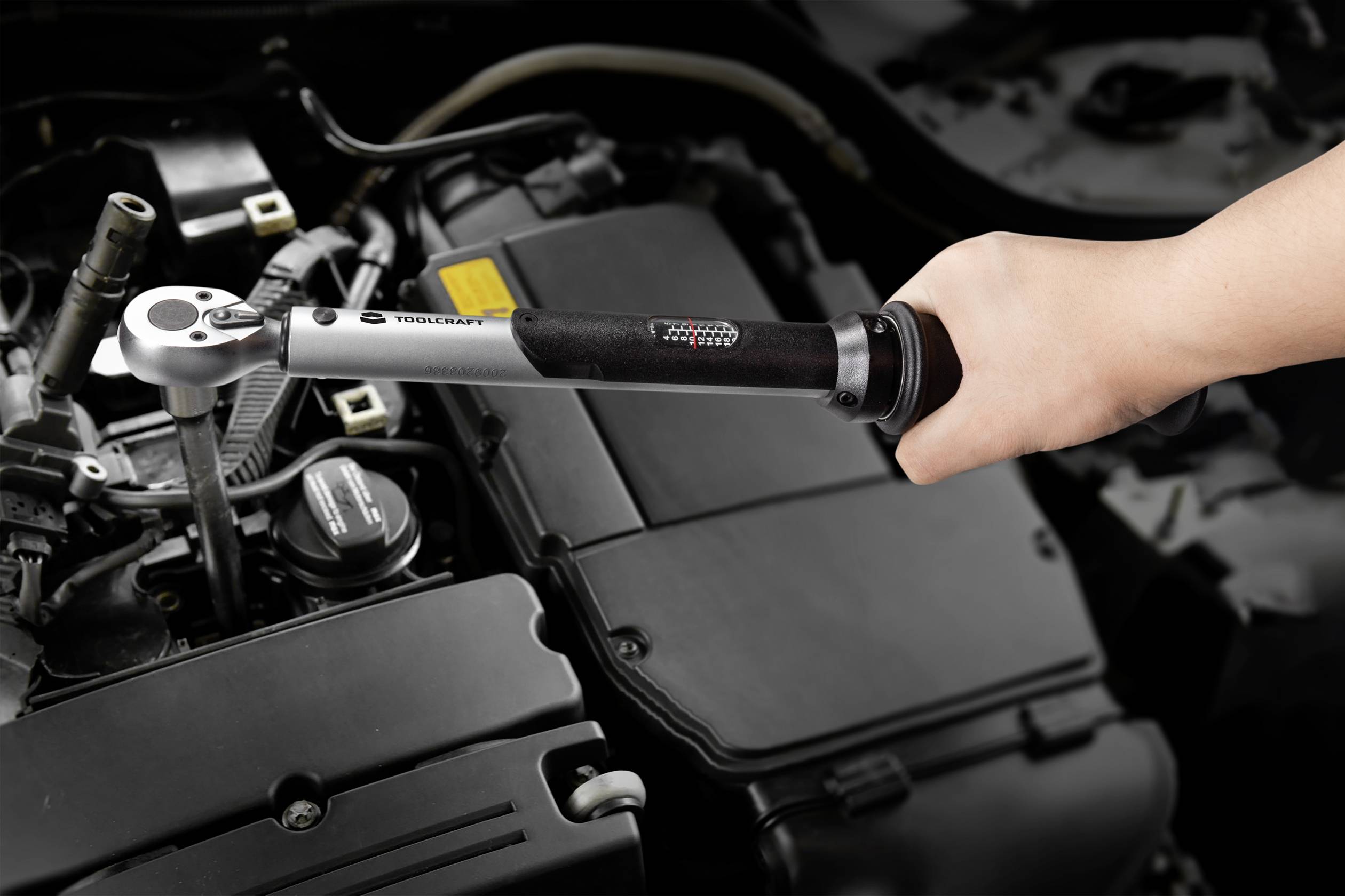 A hand is tightening a screw with a torque wrench in a car engine compartment. The engine compartment is partially visible.
