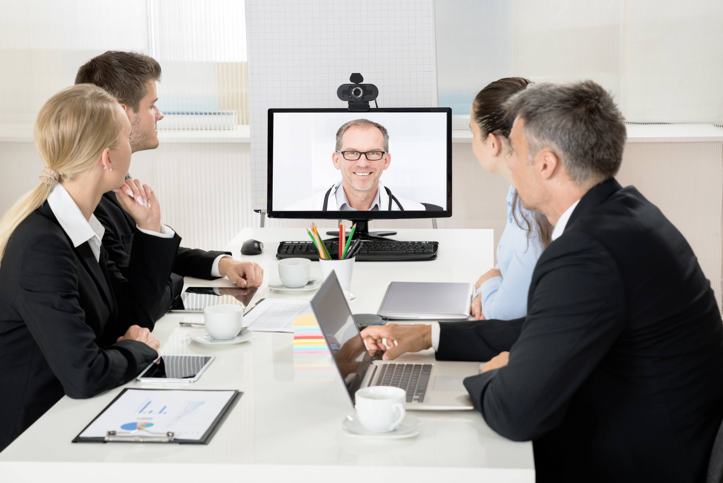 People in a meeting room look at a monitor displaying a video conference participant. Laptops and notes are on the table.