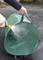 A person is spraying water from a garden hose onto a large, green, empty carrier bag outdoors on an asphalt surface.