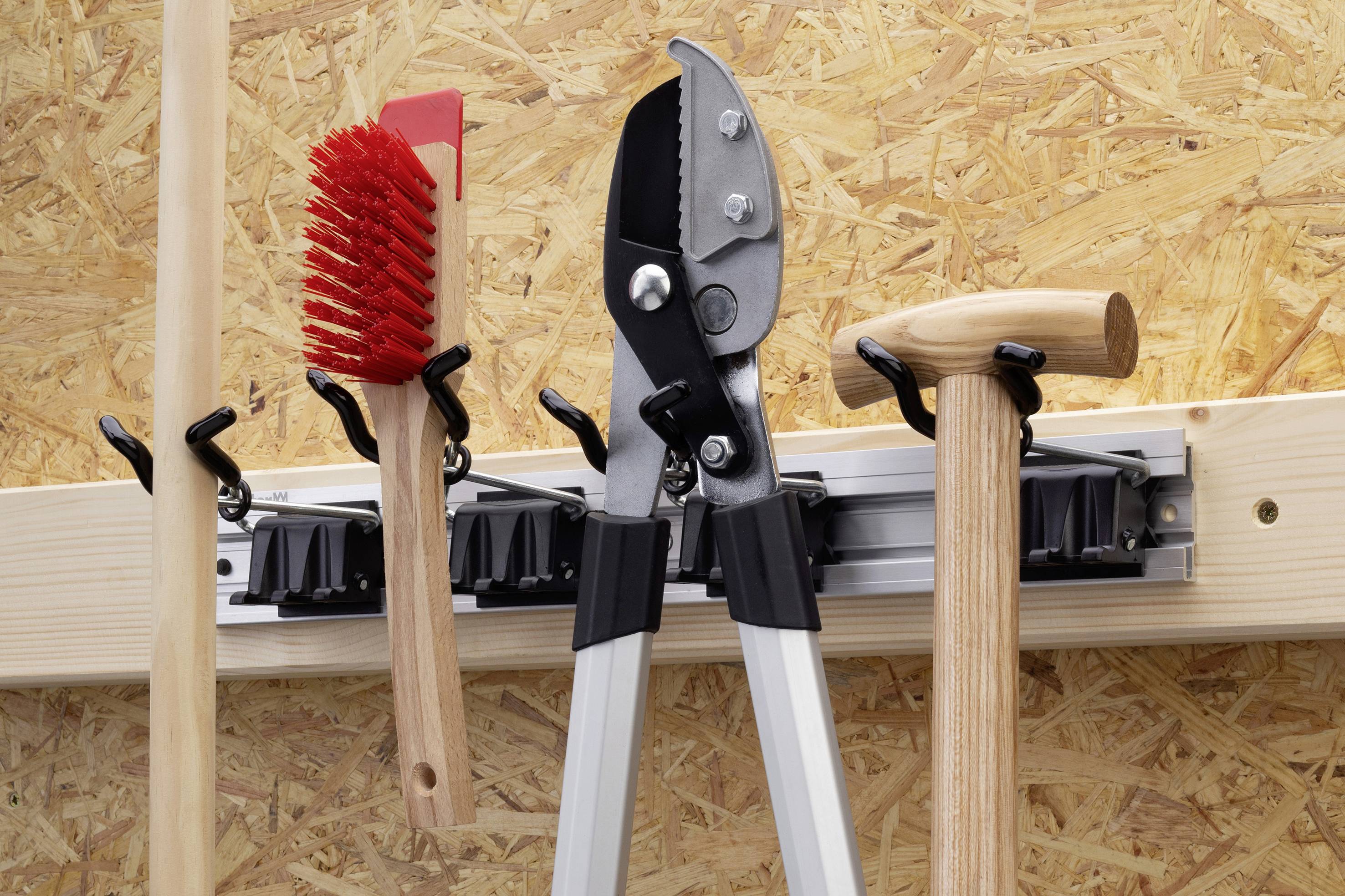 Tools on a wooden wall: a red broom, a pair of pruning shears, and a hammer hanging on a wall mounting system.