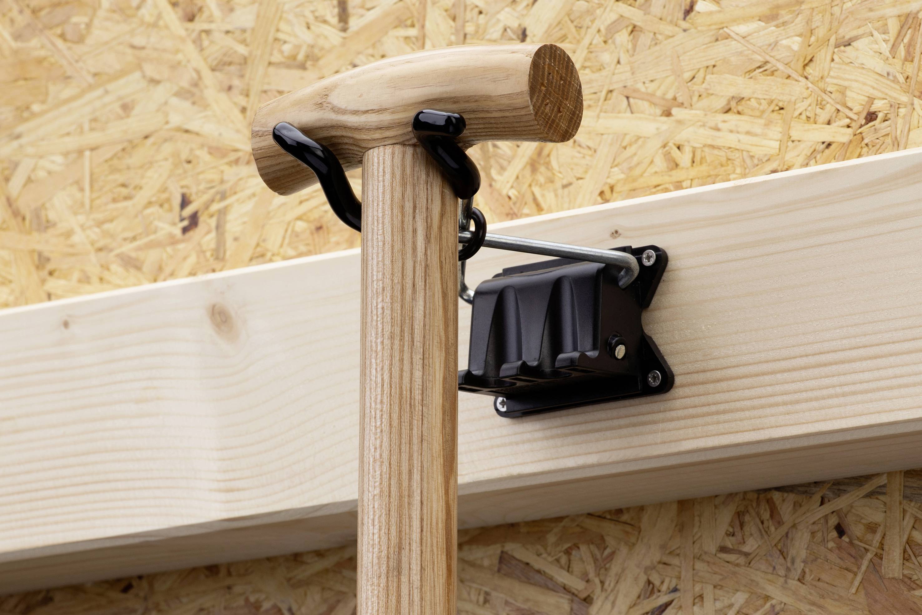 Wooden handle of a tool hanging on a metal wall hook bracket, mounted on a wooden board with a chipboard background.