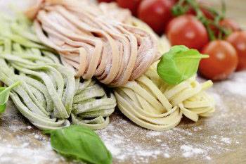 Colourful pasta in spinach, tomato, and plain flavours lie side by side on a floured board, surrounded by fresh tomatoes and basil.