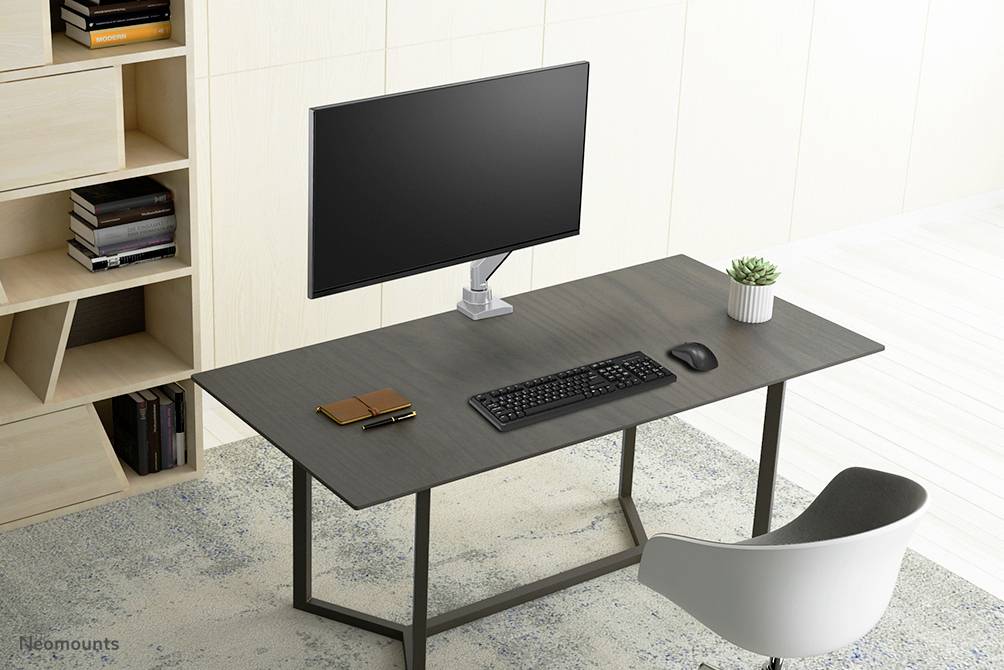 A tidy desk with a monitor, keyboard, mouse and a notebook. In the background, a bookshelf with books; a modern, minimalist interior.