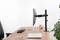 Monitor arm on a desk, holding a screen. Laptop, keyboard, mouse, clock and plants in the background.